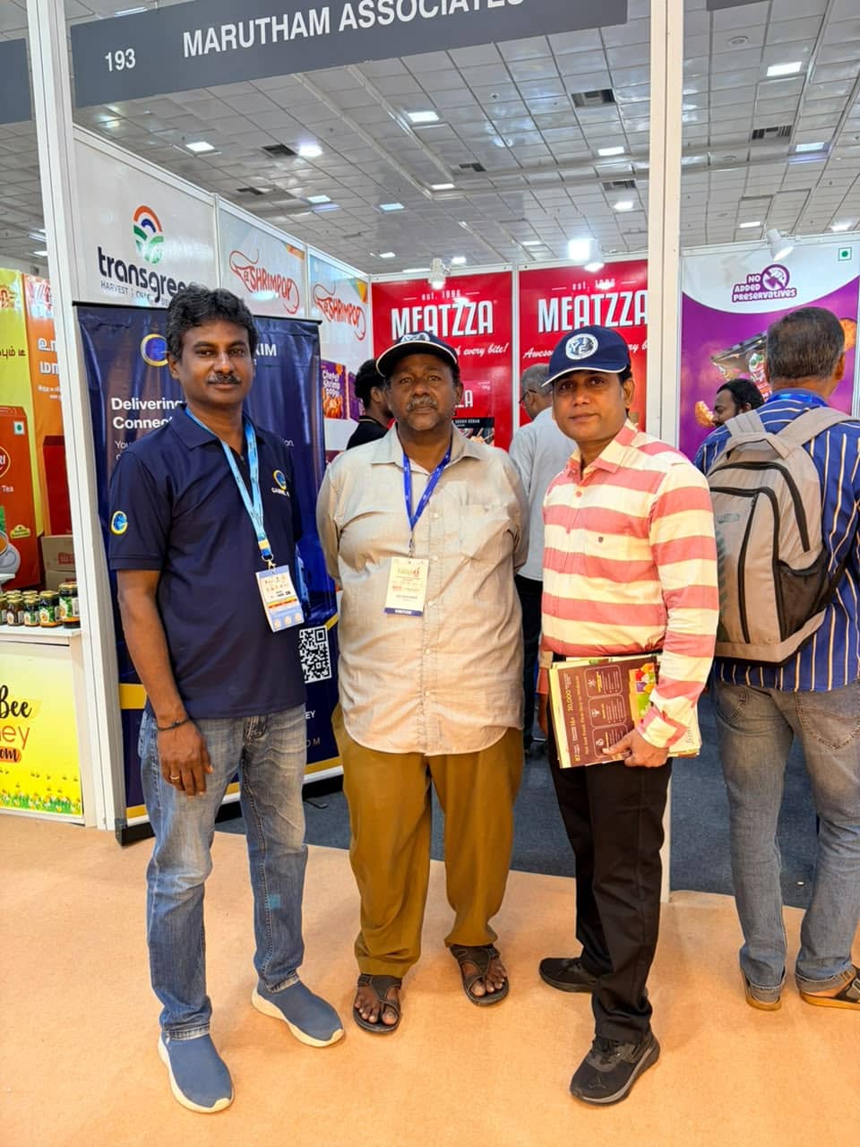 Three men posing at a trade show booth labeled Marutham Associates with red Meatzza banners behind them, badges on lanyards visible.